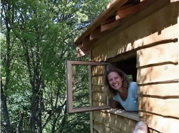 Une femme passe la tête par la fenêtre de sa cabane dans les arbres, durant son weekend nature.